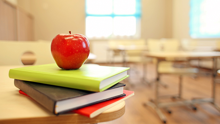 photo of stack of books with apple on top with classroom desks in background